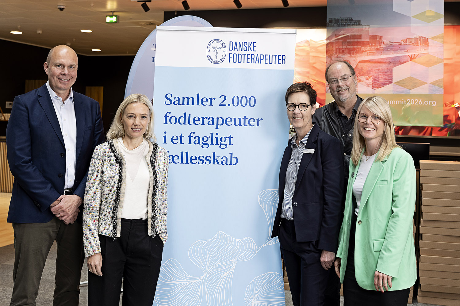 Du ser fem mennesker stå på hver sin side af en roll-up, hvor der står; Samler 2.000 fodterapeuter i et fagligt fælleskab.