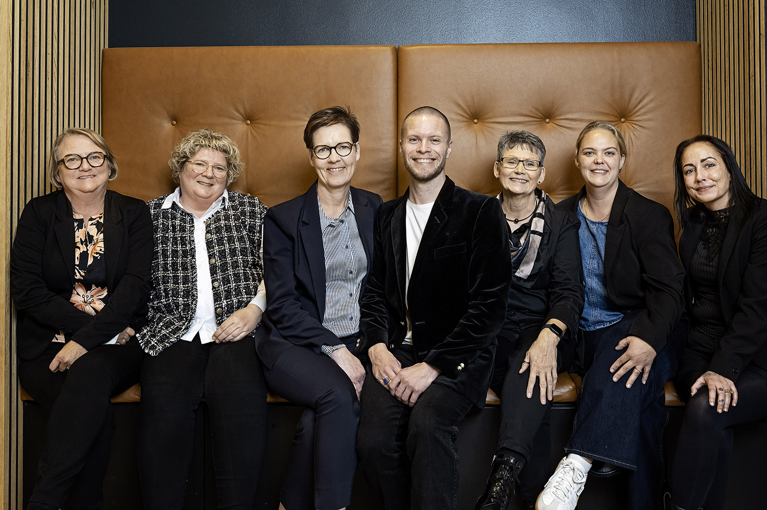 En gruppe smilende fodterapeuter sidder tæt sammen i en lysebrun lædersofa med høje ryglæn. 